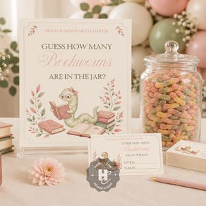 May include: A game set featuring a sign and cards that read "Guess how many Bookworms are in the jar?" with an illustration of a worm reading books, a clear glass jar filled with gummy worms, and a pink pen on a light-colored surface.