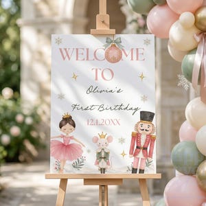 May include: A rectangular welcome sign on a wooden easel featuring the text "WELCOME TO Olivia's First Birthday 12.1.20XX". The design includes a pink ornament, a ballerina in a pink tutu, a mouse in a crown, and a nutcracker with gold accents.