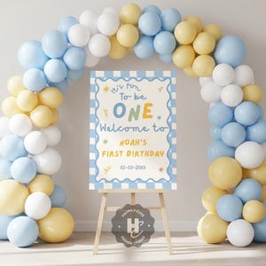 Può includere: Decorazione per festa di compleanno con un arco di palloncini nei colori pastello blu, giallo e bianco. Un cartello su un cavalletto di legno recita "It's fun to be ONE, Welcome to Noah's First Birthday" con la data 03-03-20XX.