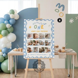 May include: A wooden easel displays a birthday sign with the text "It's fun to be ONE" and "Noah's First Birthday." The sign has a blue and white checkered border and includes photos of a baby. Balloons and cupcakes are in the background.