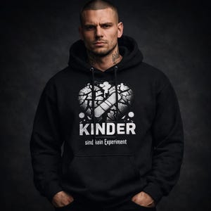 Puede incluir: Sudadera con capucha negra con un diseño gráfico de un corazón roto con una tirita y la palabra "KINDER" en blanco. El texto "sind kein Experiment" está debajo de la palabra "KINDER".