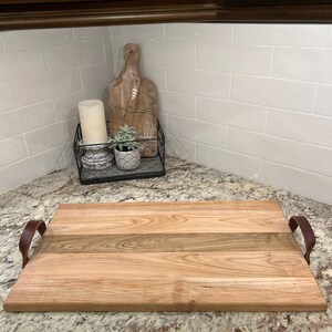May include: A large wooden serving tray with two leather handles. The tray is made of light and dark wood with a natural finish. The tray is sitting on a granite countertop.