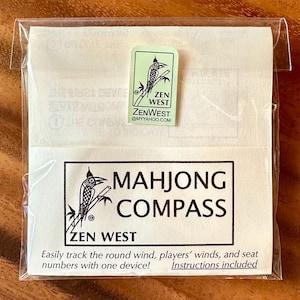 Peut inclure: Un emballage plastique transparent contenant un Mahjong Compass. L'emballage comporte un autocollant vert avec une illustration d'oiseau et le texte "ZEN WEST". L'&eacute;tiquette principale indique "MAHJONG COMPASS" avec "ZEN WEST" en dessous. Le texte "Easily track the round wind..." est &eacute;galement visible.