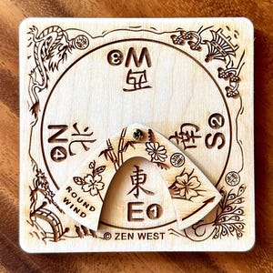 Peut inclure: Une boussole carr&eacute;e en bois avec des motifs et du texte grav&eacute;s d'inspiration asiatique. La boussole comporte un pointeur rotatif et est &eacute;tiquet&eacute;e avec les points cardinaux et le texte "ROUND WIND" et "ZEN WEST".