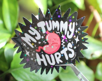 May include: A black sticker with a starburst design and the words "MY TUMMY HURTS" in white. A pink, cartoon stomach with a sad face is in the center. The sticker has holographic accents and is held by tweezers.