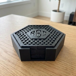 May include: Black hexagonal coaster set with a matching holder. The coasters have a textured surface with a repeating pattern of small circles. The center of the top coaster features the letters "RED".