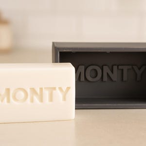 May include: A rectangular bar of off-white soap with the name "MONTY" embossed on the front, next to a dark gray rectangular soap dish with the same name. The soap and dish are on a light-colored surface.