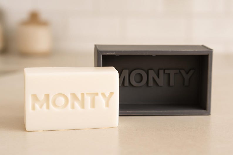 May include: A rectangular bar of off-white soap with the name "MONTY" embossed on the front, next to a dark gray rectangular soap dish with the same name. The soap and dish are on a light-colored surface.