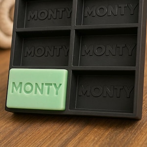 May include: A black soap dish with six compartments, each labeled "MONTY" in raised letters. A light green soap bar with "MONTY" is in one compartment. The dish sits on a wooden surface.