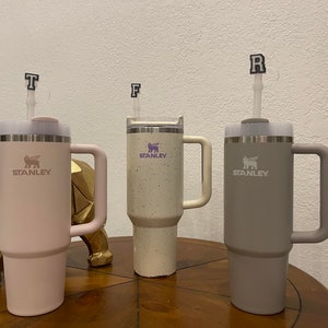 May include: Three stainless steel travel mugs with handles. The mugs are in pink, white with black speckles, and gray. Each mug has a logo that says "STANLEY" and a small animal graphic. The mugs have lids with straws and letter charms on top.
