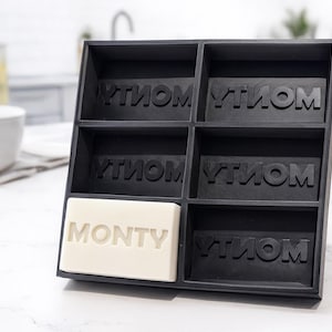 May include: A black soap holder with six compartments, each labeled with the word "MONTY" in a repeating pattern. A single bar of white soap, also labeled "MONTY", sits in one compartment. The holder is on a white surface.