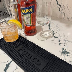 May include: Aperol bottle, cocktail shaker, and a wine glass on a countertop. A cocktail with ice and a lemon wedge sits in a rocks glass. A black bar mat with the word "Ponto" is in the foreground.
