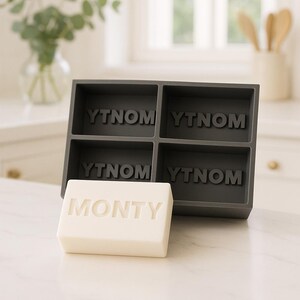 May include: A dark gray soap mold with four compartments, each labeled "YTNO M". A white bar of soap with "MONTY" engraved on it sits in front of the mold. The scene is set in a bright kitchen.