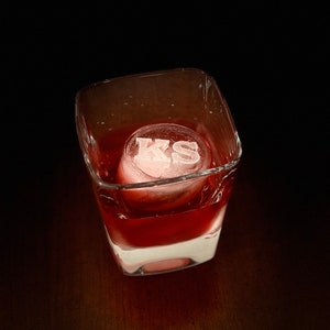 May include: A clear glass with a red liquid and a white ice cube with the letters "KS" on it.