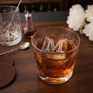 May include: A rocks glass with a diamond pattern contains a large ice cube with the letter "M" inside. The glass sits on a wooden surface, with a cocktail spoon, a mixing glass, and a small bottle in the background. The drink is amber-colored.