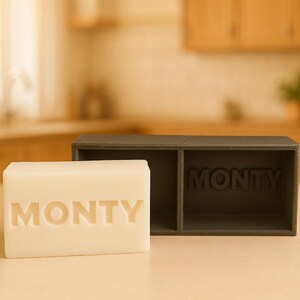 May include: A rectangular bar of white soap with the name "MONTY" carved into it, next to a dark gray soap dish with the name "MONTY" inside. The items are on a light-colored surface.