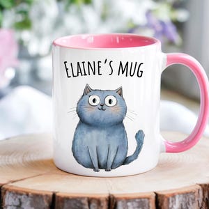 Puede incluir: Taza de cerámica blanca con interior y asa rosas. La taza presenta una ilustración de gato azul y el texto "Elaine's Mug" en negro. La taza está sobre una superficie de madera.