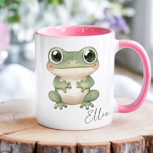 May include: White ceramic mug with a pink interior and handle. The mug features a cute cartoon frog illustration with large eyes. The name "Ellie" is written in cursive below the frog. The mug is sitting on a wooden surface.
