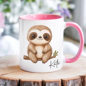 Peut inclure: Un mug en céramique blanc avec un intérieur et une anse roses. L'avant du mug présente un paresseux brun de dessin animé assis sur une branche avec le prénom Kate écrit en écriture cursive noire sous l'illustration du paresseux.