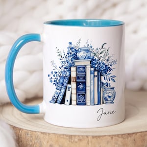 Op de afbeelding: Een witte keramische mok met een felblauw handvat en een blauwe binnenrand. De buitenkant is voorzien van een bedrukte illustratie van een stapel blauwe en witte boeken, omringd door blauwe bloemstukken en twee kleine vazen. De naam Jane staat in het zwart.