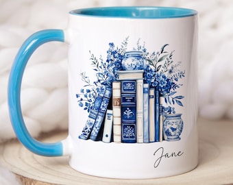 May include: A white ceramic mug with a bright blue handle and interior rim. The exterior features a printed illustration of a stack of blue and white books surrounded by blue floral arrangements and two small vases. The name Jane is printed in black.