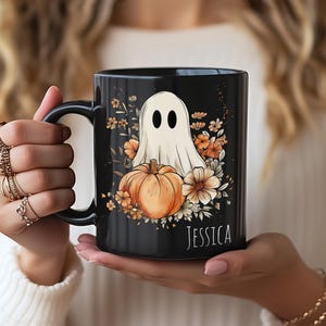 May include: A black ceramic mug featuring a white ghost illustration sitting behind an orange pumpkin surrounded by autumn flowers. The name JESSICA is printed in white capital letters at the bottom right of the mug design.