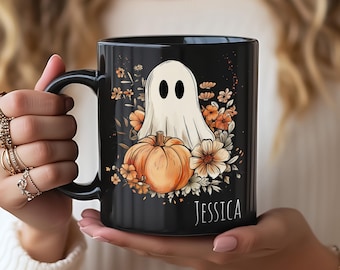 Op de afbeelding: Een zwarte keramische mok met een witte geestillustratie die achter een oranje pompoen zit, omringd door herfstbloemen. De naam JESSICA staat in witte hoofdletters rechtsonder op het ontwerp gedrukt.