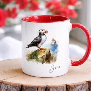 Puede incluir: Una taza de cerámica blanca con asa e interior de color rojo brillante. El exterior presenta una ilustración en acuarela de un frailecillo sobre un acantilado herboso frente al mar, con el nombre Dora escrito en cursiva negra debajo.