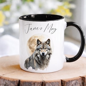 Peut inclure: Un mug en céramique blanche avec un intérieur et une anse noirs. L'avant présente une illustration style aquarelle d'un loup gris devant une pleine lune. Au-dessus du loup, le texte "Jane's Mug" est imprimé en police cursive noire.