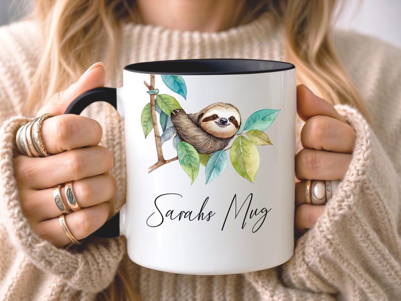 May include: White ceramic mug with a black interior and handle. The mug features a watercolor illustration of a sloth on a branch with green leaves. The text "Sarahs Mug" is written in cursive below the sloth illustration.
