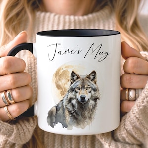 Peut inclure: Un mug en céramique blanc avec une anse et un bord noirs, orné d'une illustration style aquarelle représentant un loup gris devant une pleine lune. Le texte "Jane's Mug" est imprimé en police cursive noire au-dessus du motif du loup.