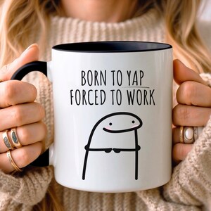 May include: A white ceramic mug with a black handle and interior rim. The mug features the black text BORN TO YAP FORCED TO WORK above a simple black line drawing of a cartoon character with pink cheeks and hands clasped together in front.