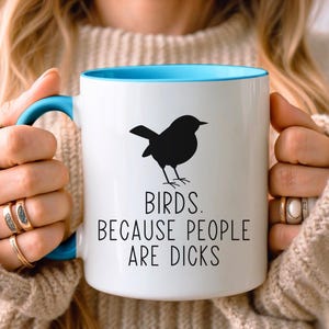 Pode incluir: Caneca de cer&acirc;mica branca com interior e al&ccedil;a azuis. A caneca apresenta uma silhueta preta de um p&aacute;ssaro e o texto "BIRDS. BECAUSE PEOPLE ARE DICKS" em preto, centrado na frente.