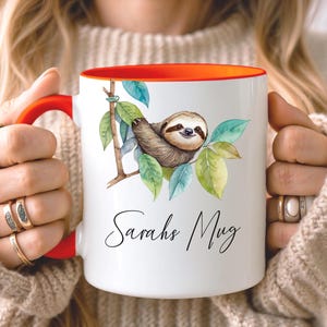 May include: White ceramic mug with a red handle and interior. The mug features a watercolor illustration of a sloth on a branch with green leaves. The text "Sarahs Mug" is written in cursive.