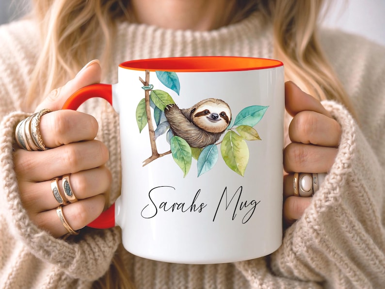 May include: White ceramic mug with a red handle and interior. The mug features a watercolor illustration of a sloth on a branch with green leaves. The text "Sarahs Mug" is written in cursive.
