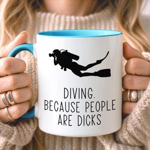 Może przedstawiać: Biały ceramiczny kubek z jasnoniebieskim wnętrzem i uchwytem. Z przodu znajduje się czarna sylwetka nurka oraz czarny napis: DIVING. BECAUSE PEOPLE ARE DICKS. Kubek ma gładkie wykończenie i standardowy cylindryczny kształt.