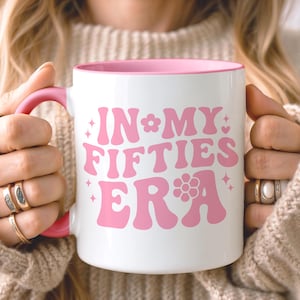 May include: A white ceramic mug with a pink interior and a pink handle. The front of the mug features the pink text "IN MY FIFTIES ERA" in a bold, wavy font, accented with small pink flower and star icons on a clean white background surface.