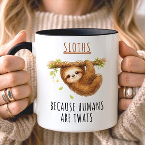 May include: A white ceramic mug with a black handle and rim, featuring a brown sloth hanging from a green branch illustration. The text SLOTHS is printed above the sloth, and the text BECAUSE HUMANS ARE TWATS is printed below the illustration.