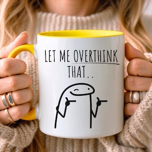 Puede incluir: Una taza de cerámica blanca con interior y asa de color amarillo brillante. El frente presenta el texto negro "LET ME OVERTHINK THAT.." sobre un dibujo lineal negro de un personaje de palitos guiñando un ojo y señalando con los dedos.