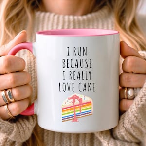 May include: White ceramic mug with a pink rim and handle. The mug has a black text that reads "I run because I really love cake" and a colorful cake illustration.