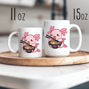 May include: Two white ceramic mugs featuring a cartoon pink axolotl eating ramen with chopsticks. The smaller mug is labelled 11 oz and the larger mug is labelled 15 oz. Both mugs display the same graphic of the axolotl holding a black bowl of noodles.