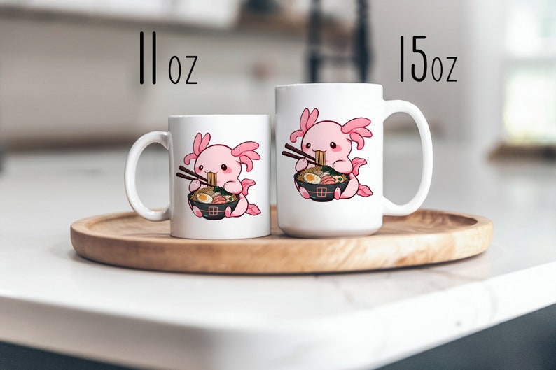 May include: Two white ceramic mugs featuring a cartoon pink axolotl eating ramen with chopsticks. The smaller mug is labelled 11 oz and the larger mug is labelled 15 oz. Both mugs display the same graphic of the axolotl holding a black bowl of noodles.