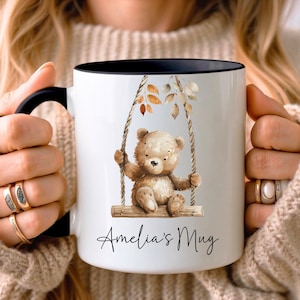 May include: White ceramic mug with a black rim. The mug features a cartoon illustration of a brown teddy bear on a swing with fall leaves. The text "Amelia's Mug" is printed on the mug.