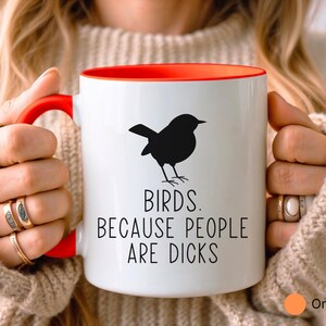 Pode incluir: Caneca de cer&acirc;mica branca com interior e al&ccedil;a vermelhos. A caneca apresenta uma silhueta preta de um p&aacute;ssaro e o texto "BIRDS. BECAUSE PEOPLE ARE DICKS" em letras pretas. Um c&iacute;rculo laranja com a palavra "Orange" est&aacute; no canto inferior direito.