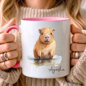 Può includere: Una tazza in ceramica bianca con interno e manico rosa, caratterizzata da un'illustrazione ad acquerello di un capibara marrone in piedi nell'acqua. Il nome Ayesha è stampato in corsivo nero sotto l'illustrazione sulla parte anteriore.