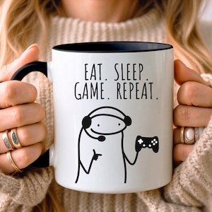 May include: A white ceramic mug with a black handle and rim, featuring the black text EAT. SLEEP. GAME. REPEAT. above a simple line drawing of a character wearing a headset and holding a game controller. The design is printed in black ink.