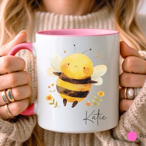 May include: White ceramic mug with a pink interior and handle. The mug features a watercolor illustration of a smiling bee with yellow and black stripes, surrounded by flowers. The name "Katie" is written in script.