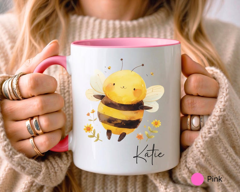 May include: White ceramic mug with a pink interior and handle. The mug features a watercolor illustration of a smiling bee with yellow and black stripes, surrounded by flowers. The name "Katie" is written in script.