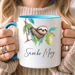 May include: White ceramic mug with a blue interior and handle. The mug features a watercolor illustration of a sloth hanging from a branch with green leaves. The text "Sarahs Mug" is written in cursive.