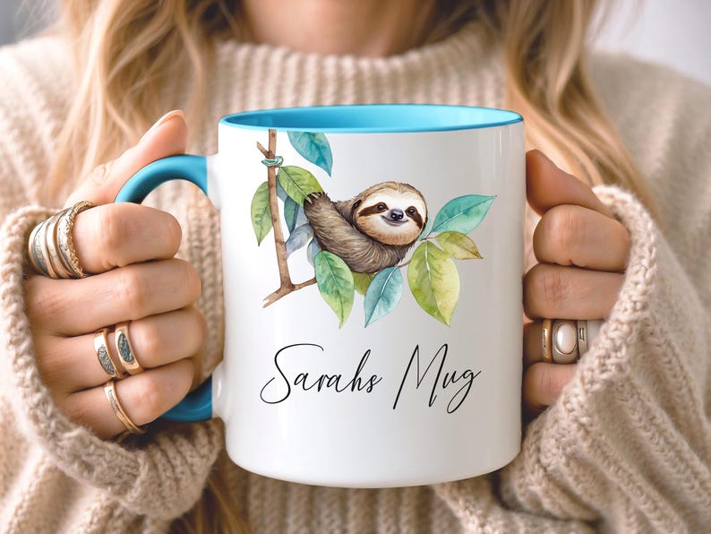May include: White ceramic mug with a blue interior and handle. The mug features a watercolor illustration of a sloth hanging from a branch with green leaves. The text "Sarahs Mug" is written in cursive.
